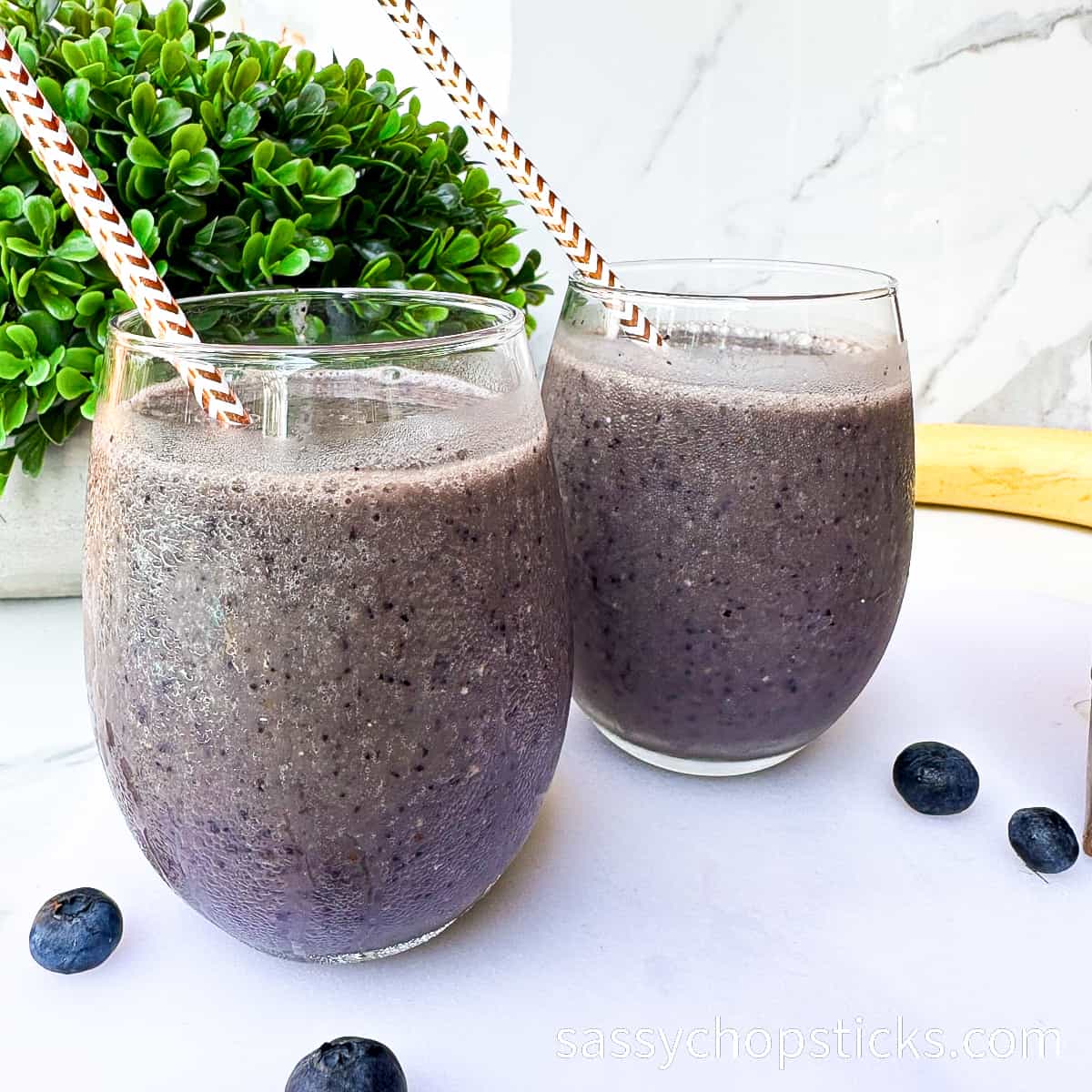 Two glasses of blueberry smoothie with straws on a white surface, garnished with a plant and scattered blueberries—one of the best drinks to brighten any moment.