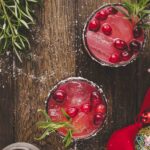 Two glasses of pink holiday cocktail with ice, cranberries, and rosemary garnish on a wooden table, surrounded by greenery, cranberries, and festive decorations.