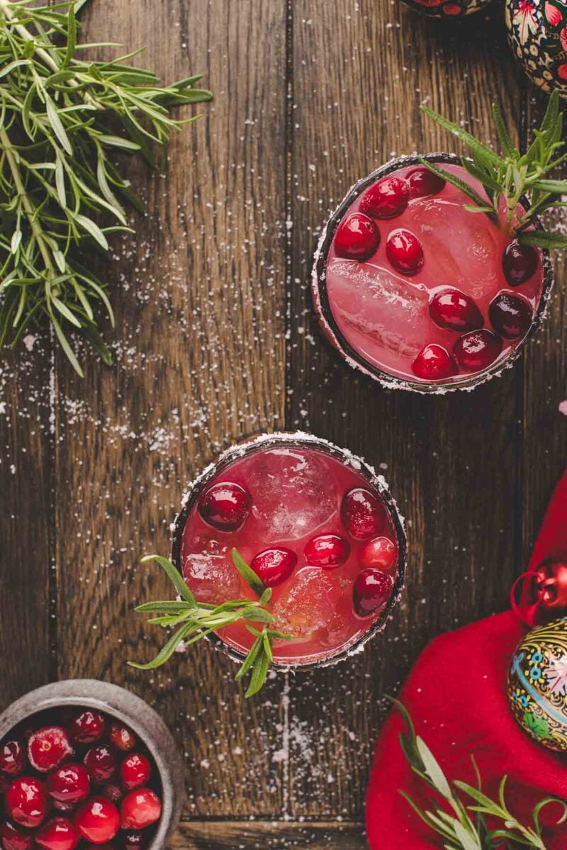Two glasses of pink holiday cocktail with ice, cranberries, and rosemary garnish on a wooden table, surrounded by greenery, cranberries, and festive decorations.