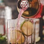 A pink cocktail is being poured from a shaker into a glass with ice, lime slices, and a salted rim.