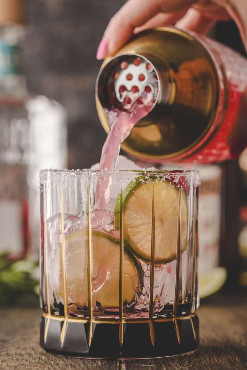 A pink cocktail is being poured from a shaker into a glass with ice, lime slices, and a salted rim.