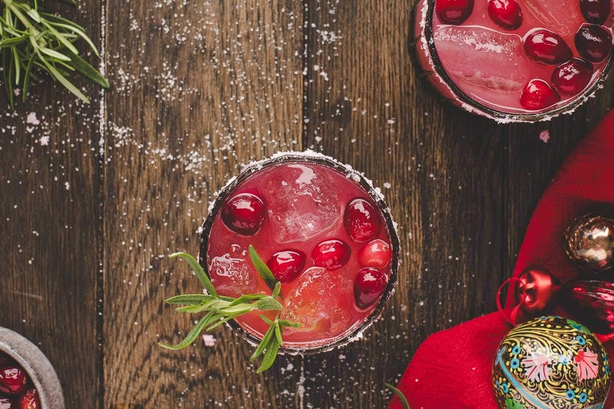 Overhead view of a cocktail with ice, cranberries, and a rosemary sprig, surrounded by holiday ornaments and a red cloth on a wooden table.