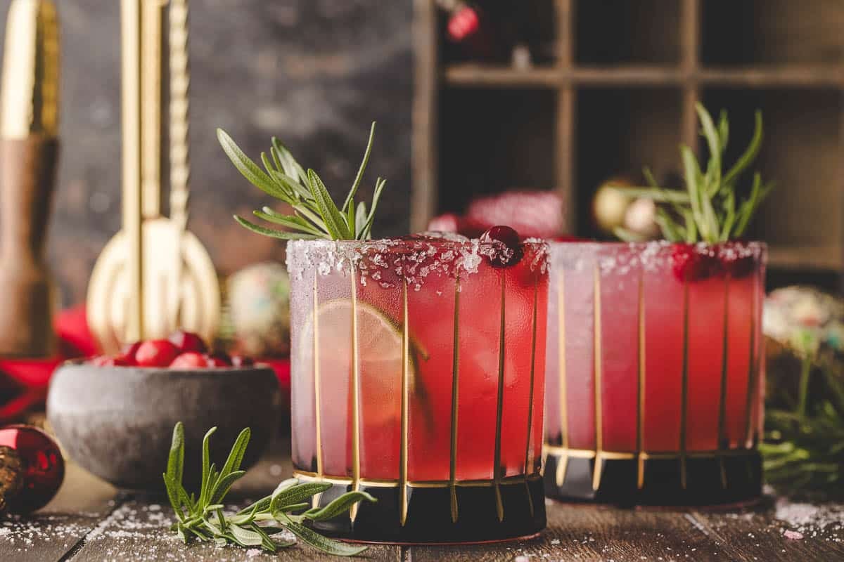 Two glasses of red cocktail garnished with lime slices, cranberries, rosemary sprigs, and salted rims on a wooden table, with bar tools and cranberries in the background.