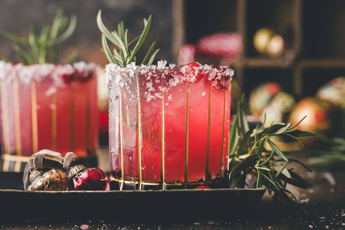 A glass of pink cocktail with a salted rim, garnished with a sprig of rosemary, sits on a tray next to holiday ornaments.
