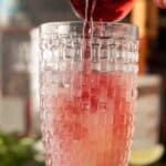 A hand pours red liquid into a tall, textured glass filled with ice and a light pink beverage.