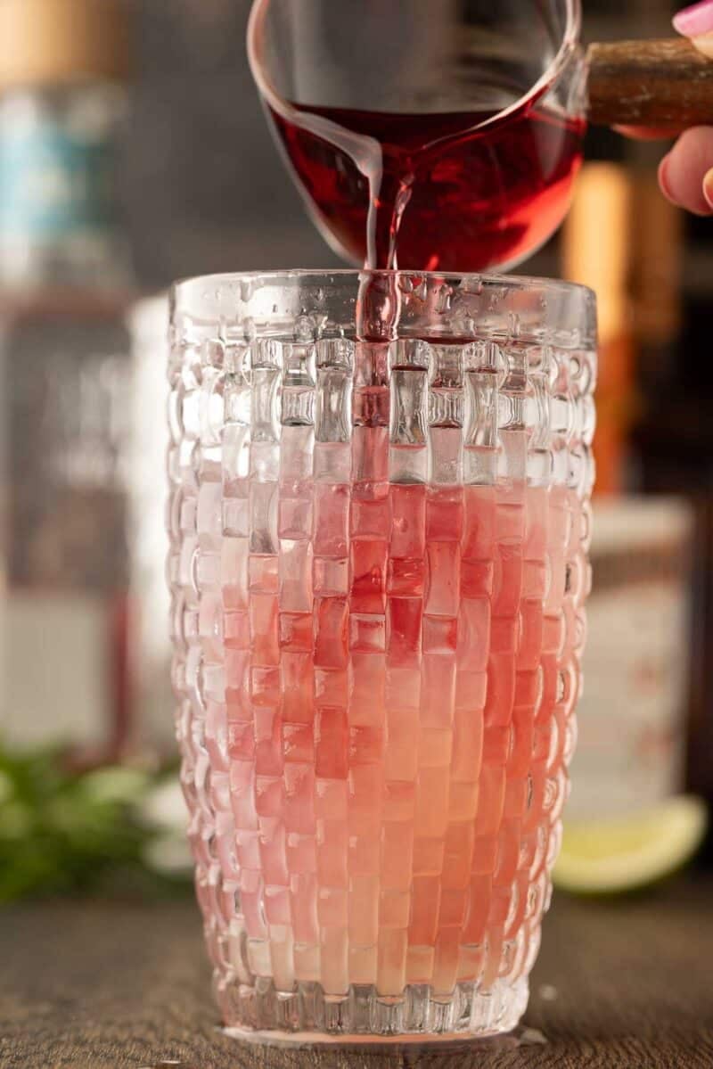 A hand pours red liquid into a tall, textured glass filled with ice and a light pink beverage.