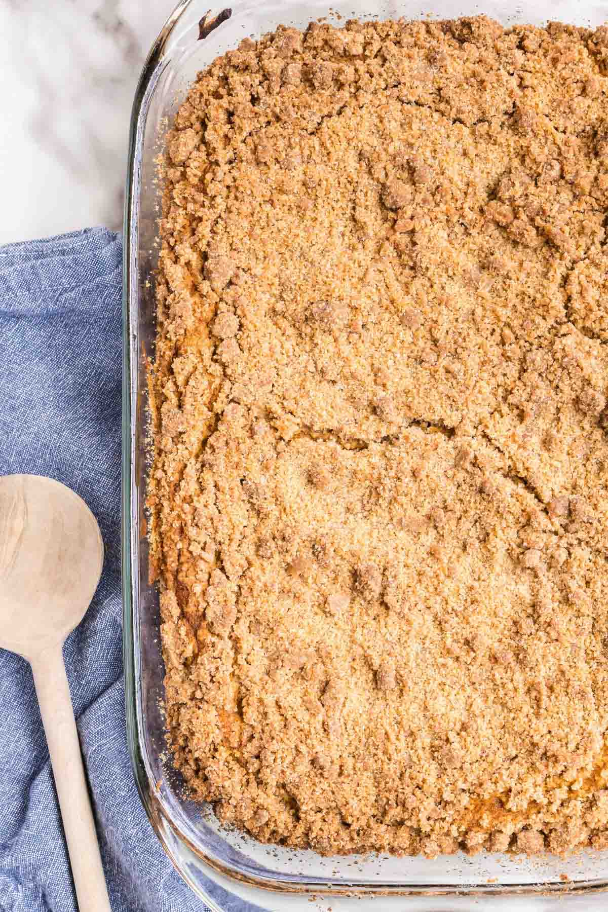 A glass baking dish filled with a golden-brown crumb-topped dessert sits next to a wooden spoon and a blue cloth.