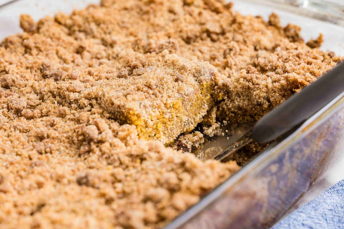 A close-up of a rectangular baking dish filled with pumpkin crumb cake, with one square piece being lifted out with a metal spatula.
