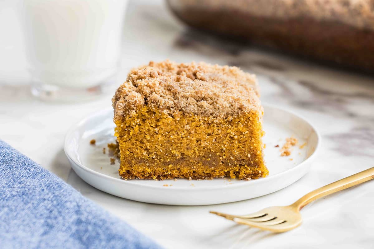 A slice of pumpkin crumb cake with a crumb topping sits on a white plate next to a gold fork, with a glass of milk and blue napkin nearby.