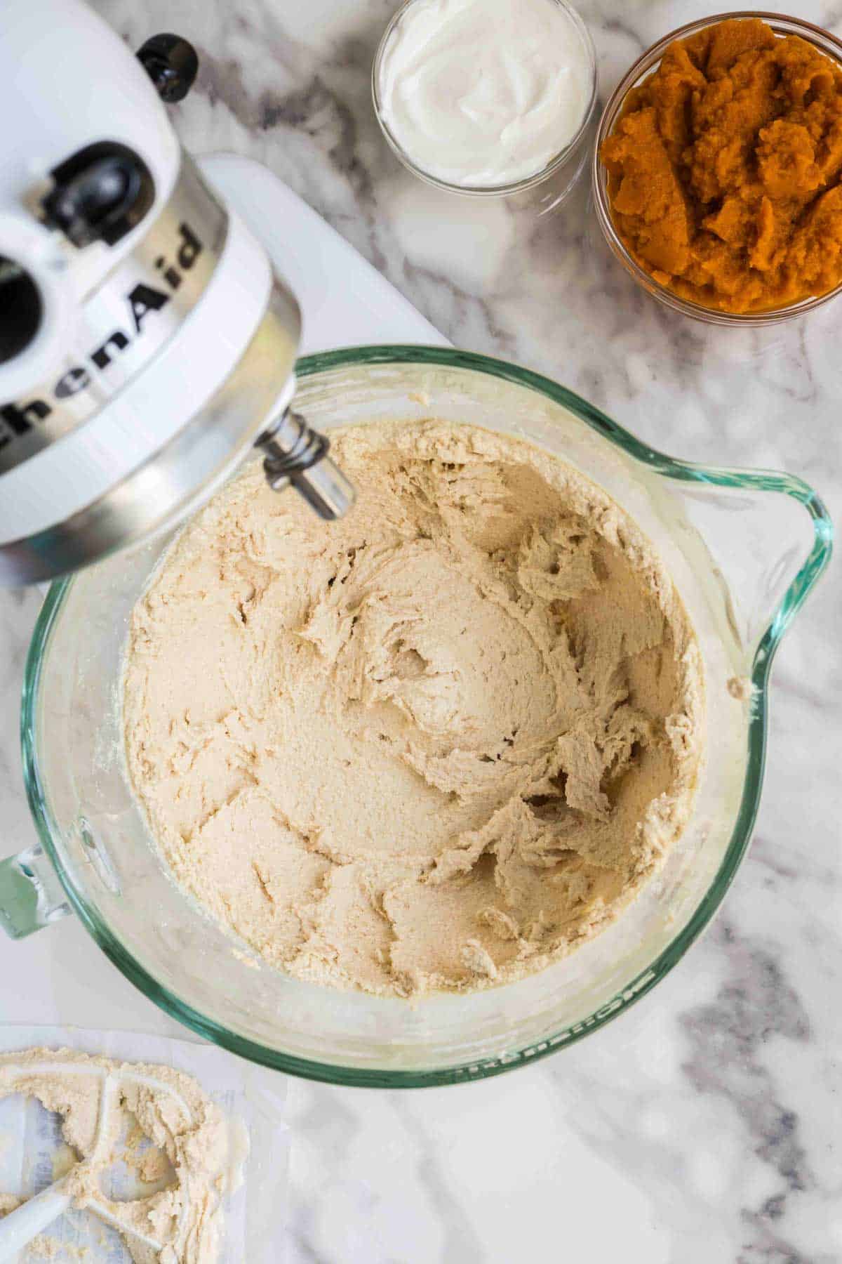 A stand mixer with creamed batter in a glass bowl, next to bowls of pumpkin puree and what appears to be sour cream on a marble countertop.