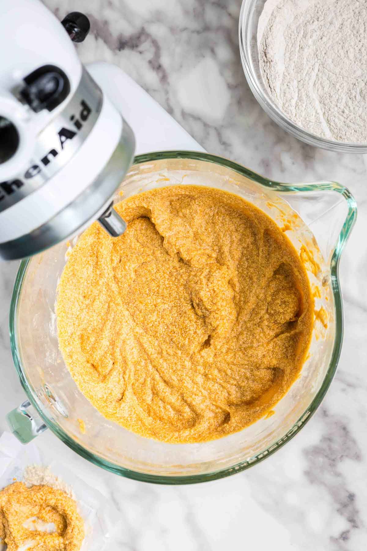 A stand mixer bowl containing a thick, yellowish batter sits on a marble countertop next to a clear bowl of dry flour mixture.