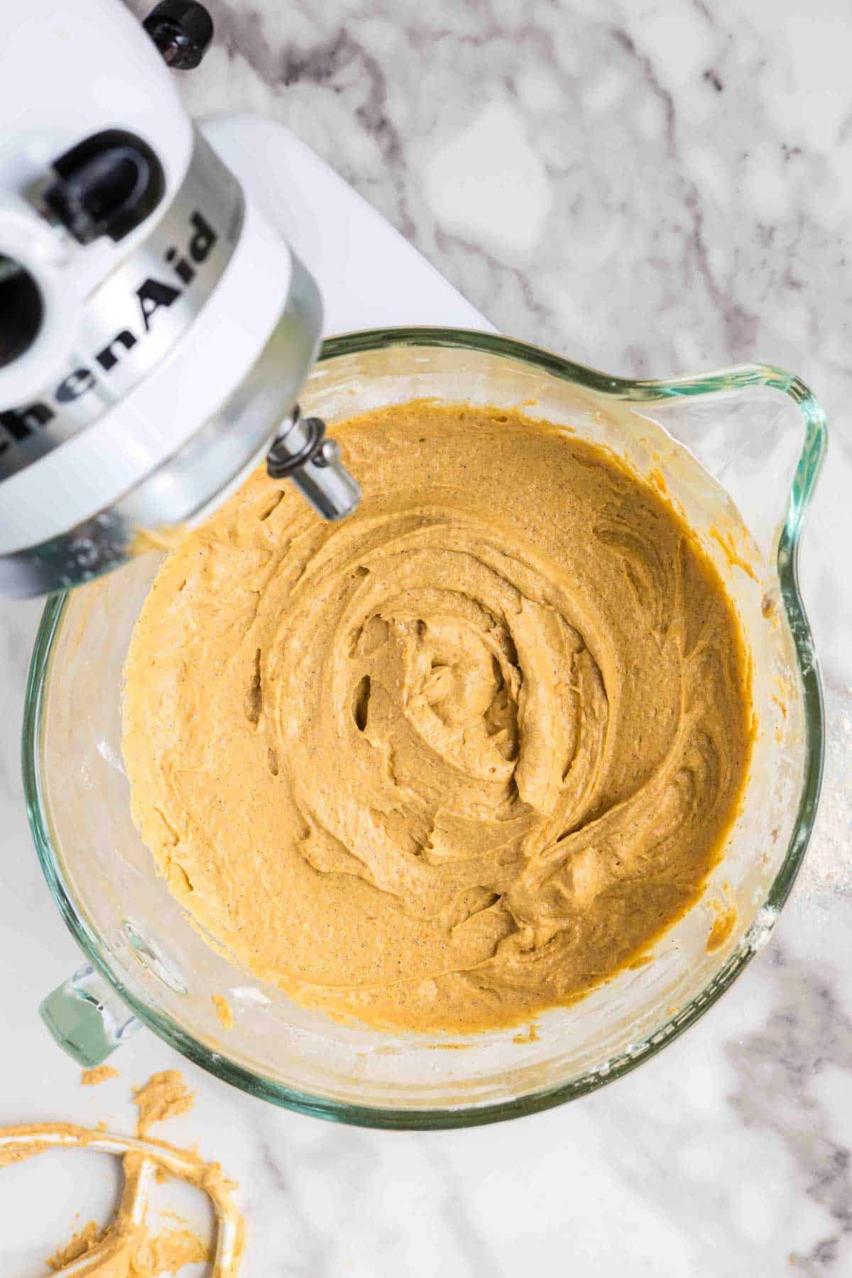 Overhead view of a glass bowl containing cookie dough being mixed with a stand mixer on a marble countertop.