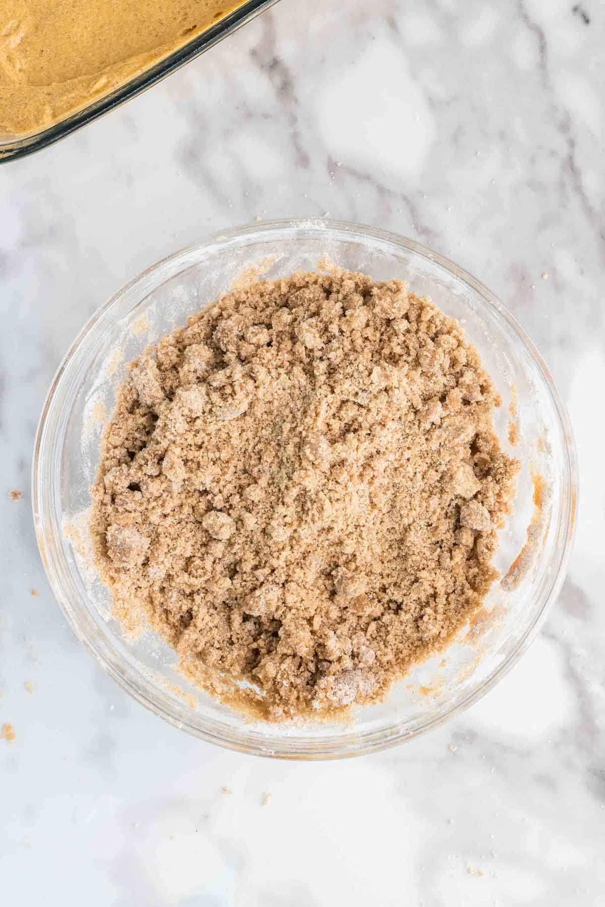 A glass bowl filled with a crumbly brown sugar and flour mixture sits on a marble countertop.