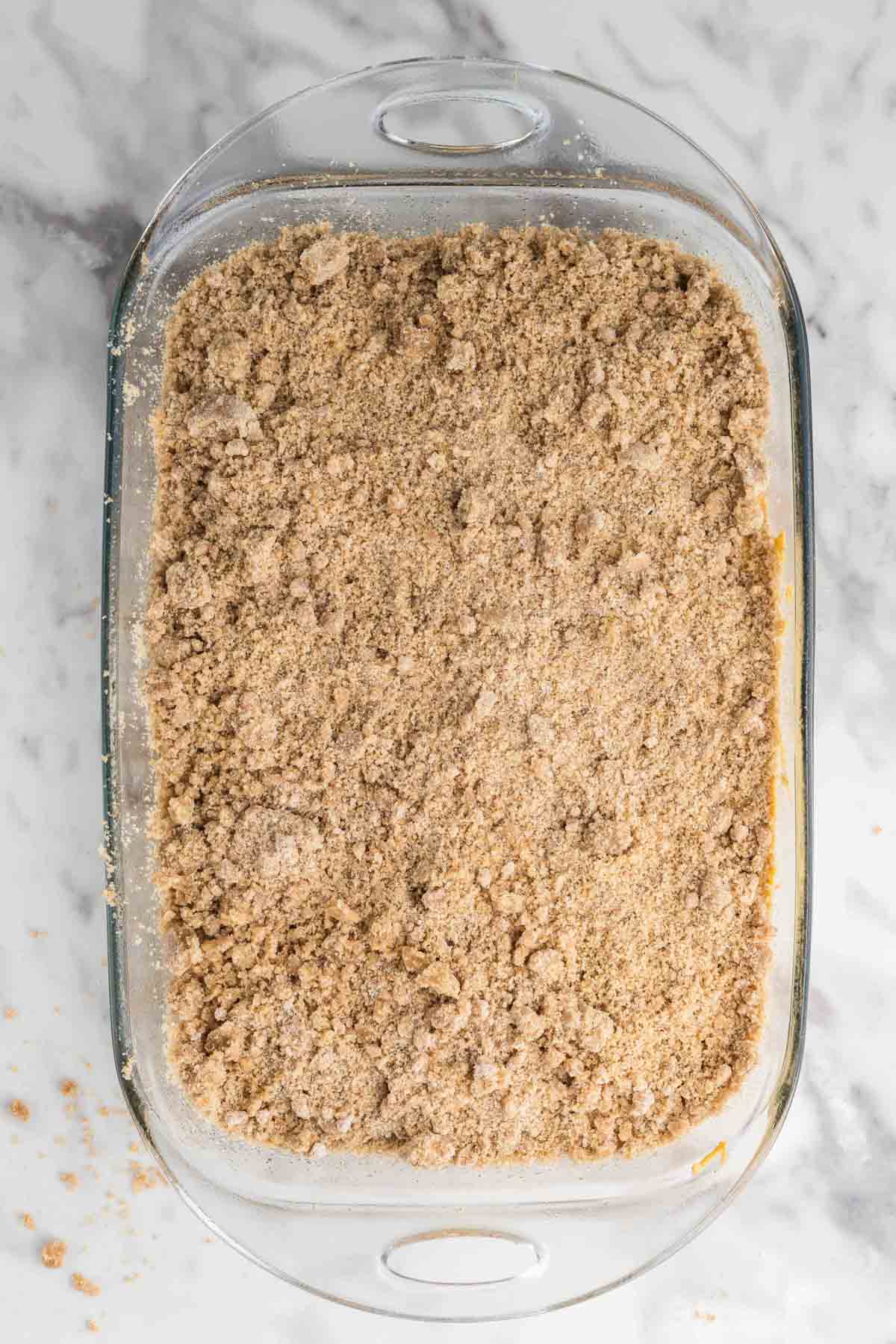 A glass baking dish filled with an unbaked crumbly cinnamon streusel topping, placed on a marble surface.