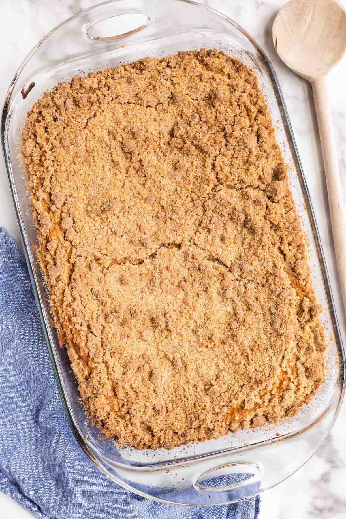 A glass baking dish filled with pumpkin crumb cake&nbsp;topped with cinnamon streusel topping, resting on a blue cloth beside a wooden spoon.