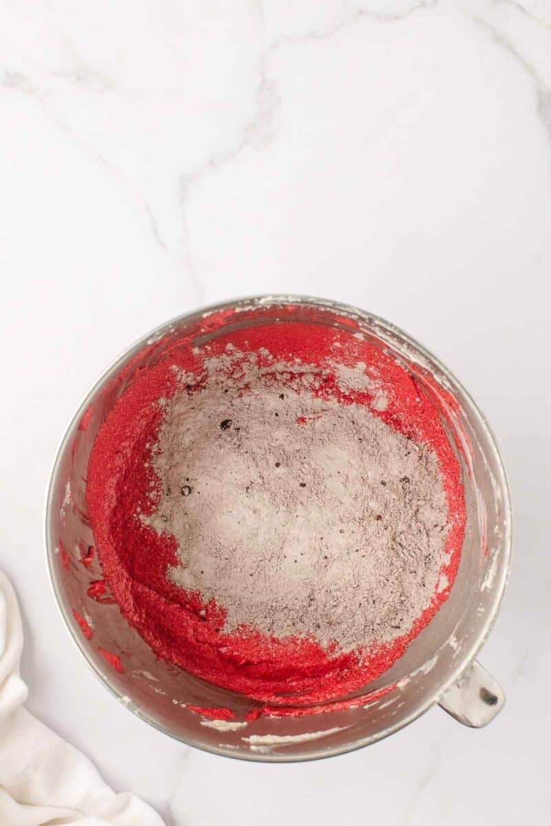 A mixing bowl containing red batter with cocoa powder and flour sitting on top, ready to be mixed, on a white marble countertop.