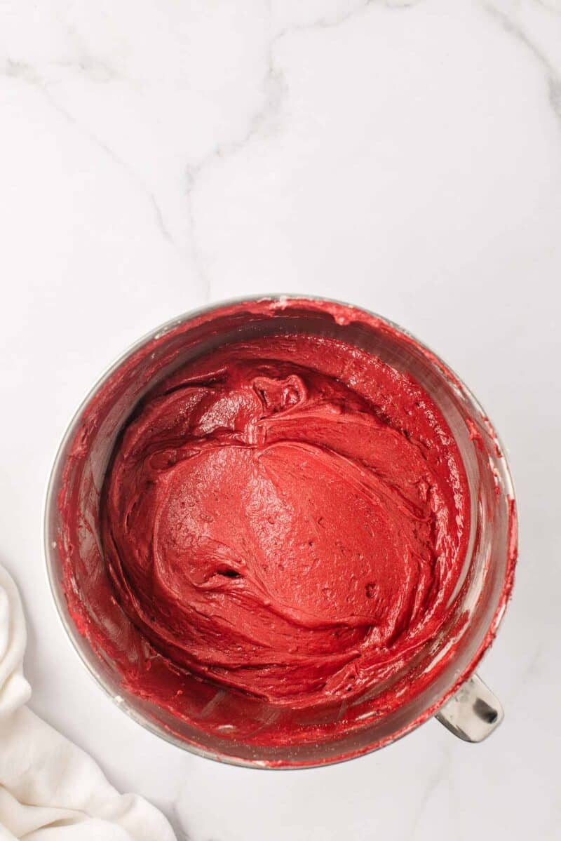 A mixing bowl filled with smooth, red cake batter sits on a white marble countertop.