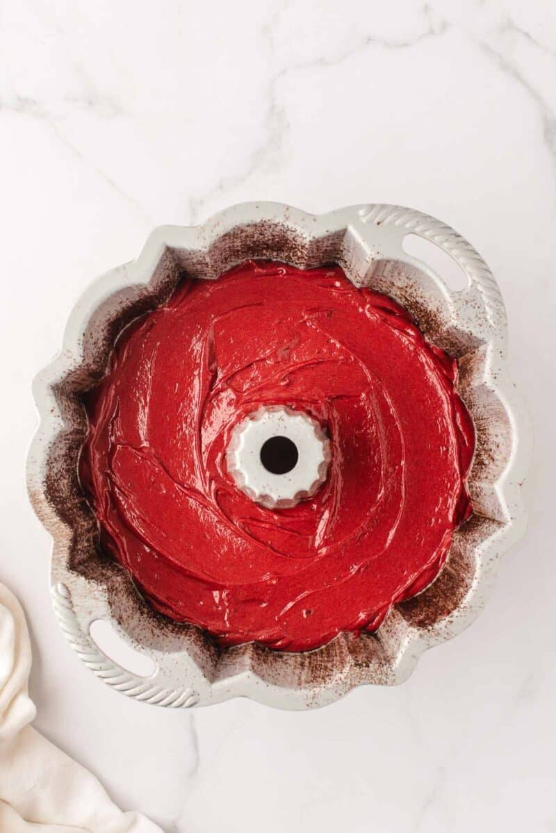 A Bundt pan filled with smooth red cake batter on a white marble surface.