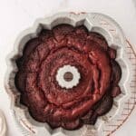 A chocolate Bundt cake in a scalloped Bundt pan resting on a round cooling rack atop a marble surface.