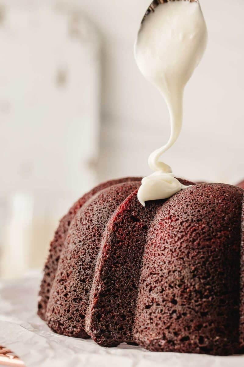 A spoon drizzles white icing onto a chocolate bundt cake sitting on parchment paper.