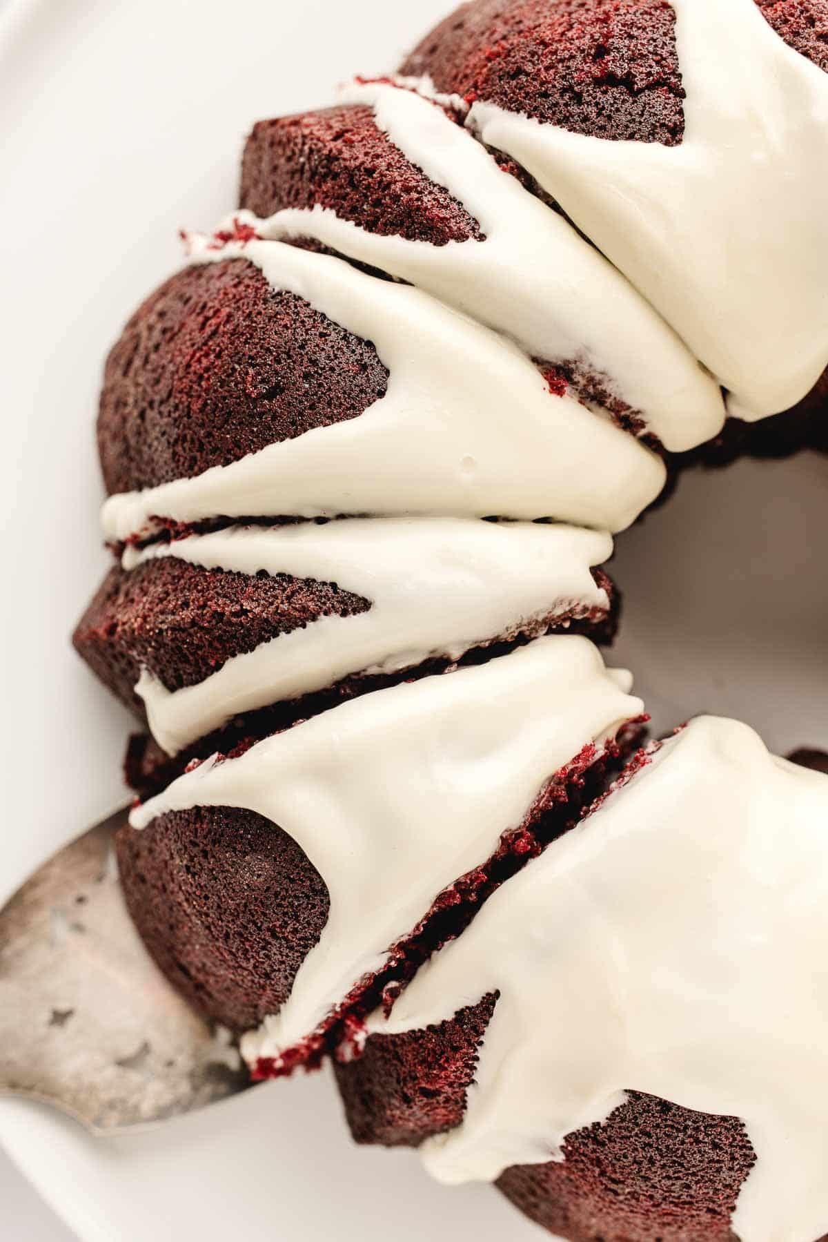 A close-up of a sliced red velvet bundt cake topped with white cream cheese icing on a white plate.