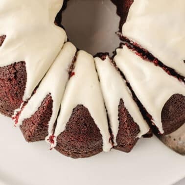 A chocolate bundt cake with white icing, partially sliced, sits on a white plate with a metal cake server.