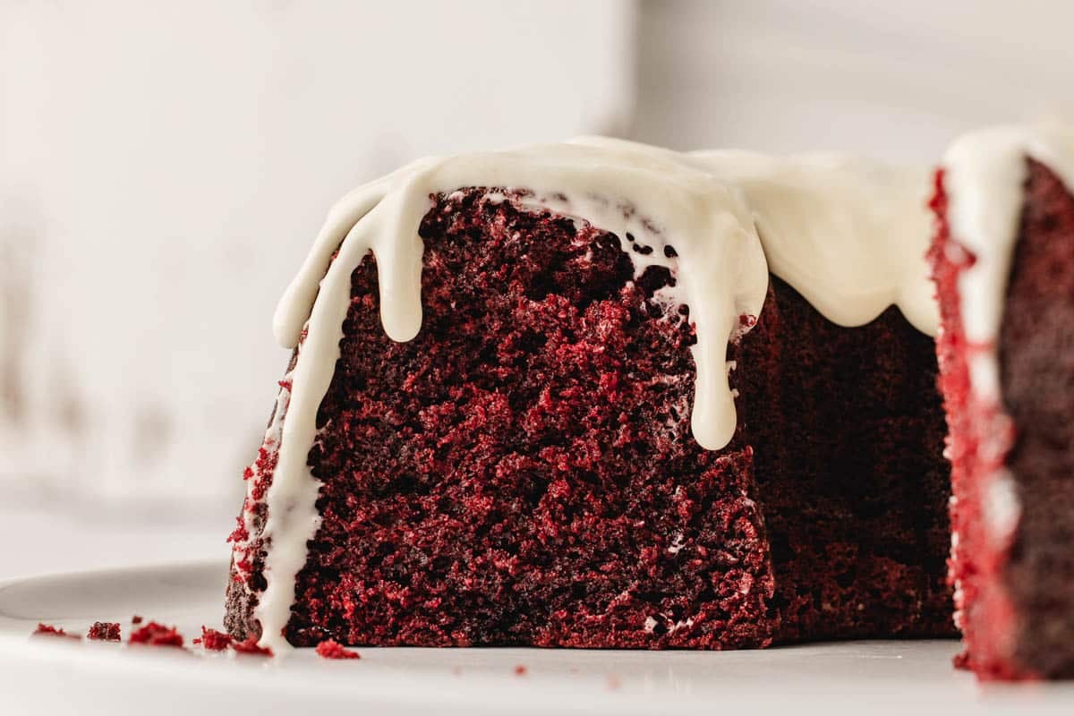 A close-up of a slice of red velvet cake with white icing dripping down the side, sitting on a white plate.