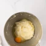 A metal mixing bowl with creamed butter and sugar, and a cracked egg added on top, sitting on a white marble countertop.
