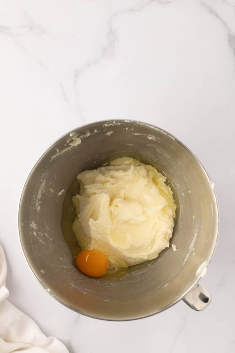 A metal mixing bowl with creamed butter and sugar, and a cracked egg added on top, sitting on a white marble countertop.