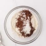A glass bowl on a marble surface containing flour, cocoa powder, baking soda, and salt, unmixed.