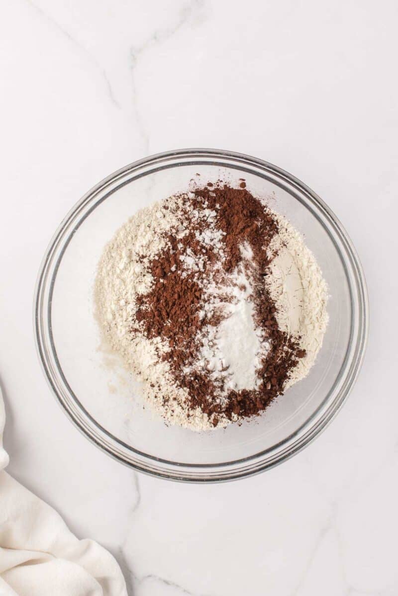 A glass bowl on a marble surface containing flour, cocoa powder, baking soda, and salt, unmixed.