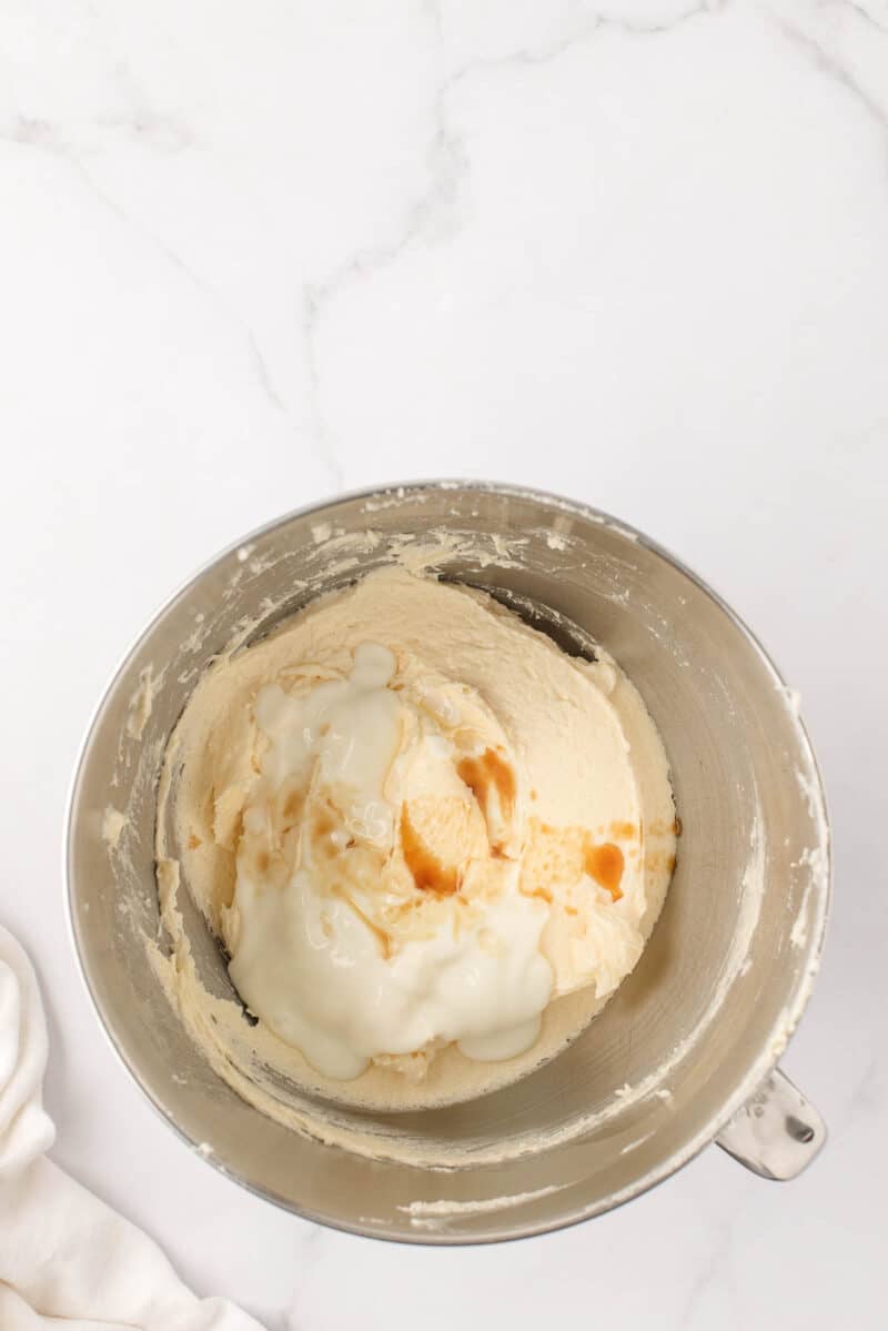 A metal mixing bowl containing a creamy batter with liquid ingredients, likely vanilla and milk, on a white marble surface.