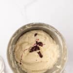 A metal mixing bowl with pale batter and a dollop of dark red jam on top, placed on a white marble surface.