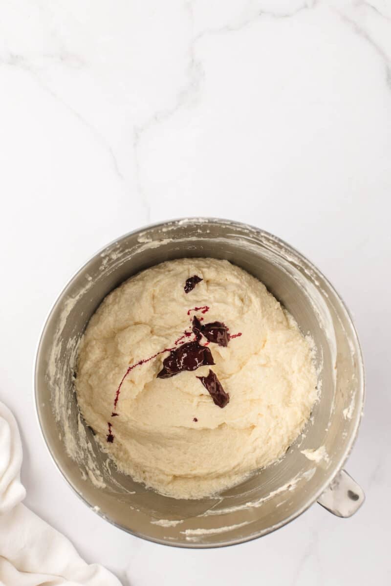 A metal mixing bowl with pale batter and a dollop of dark red jam on top, placed on a white marble surface.