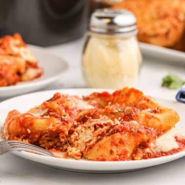 A plate of stuffed pasta shells with tomato sauce and cheese sits on a white table with a fork, a shaker of grated cheese, and garlic bread in the background.
