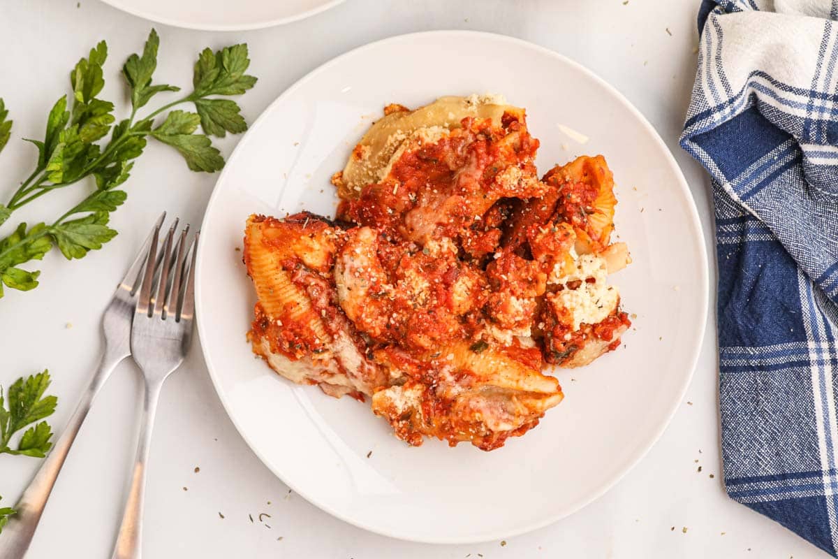A serving of stuffed pasta shells topped with tomato sauce and grated cheese on a white plate, with two forks, parsley, and a blue-and-white napkin nearby.