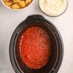 A slow cooker with tomato sauce, a bowl of uncooked pasta shells, and a bowl of ricotta cheese mixture on a white surface.
