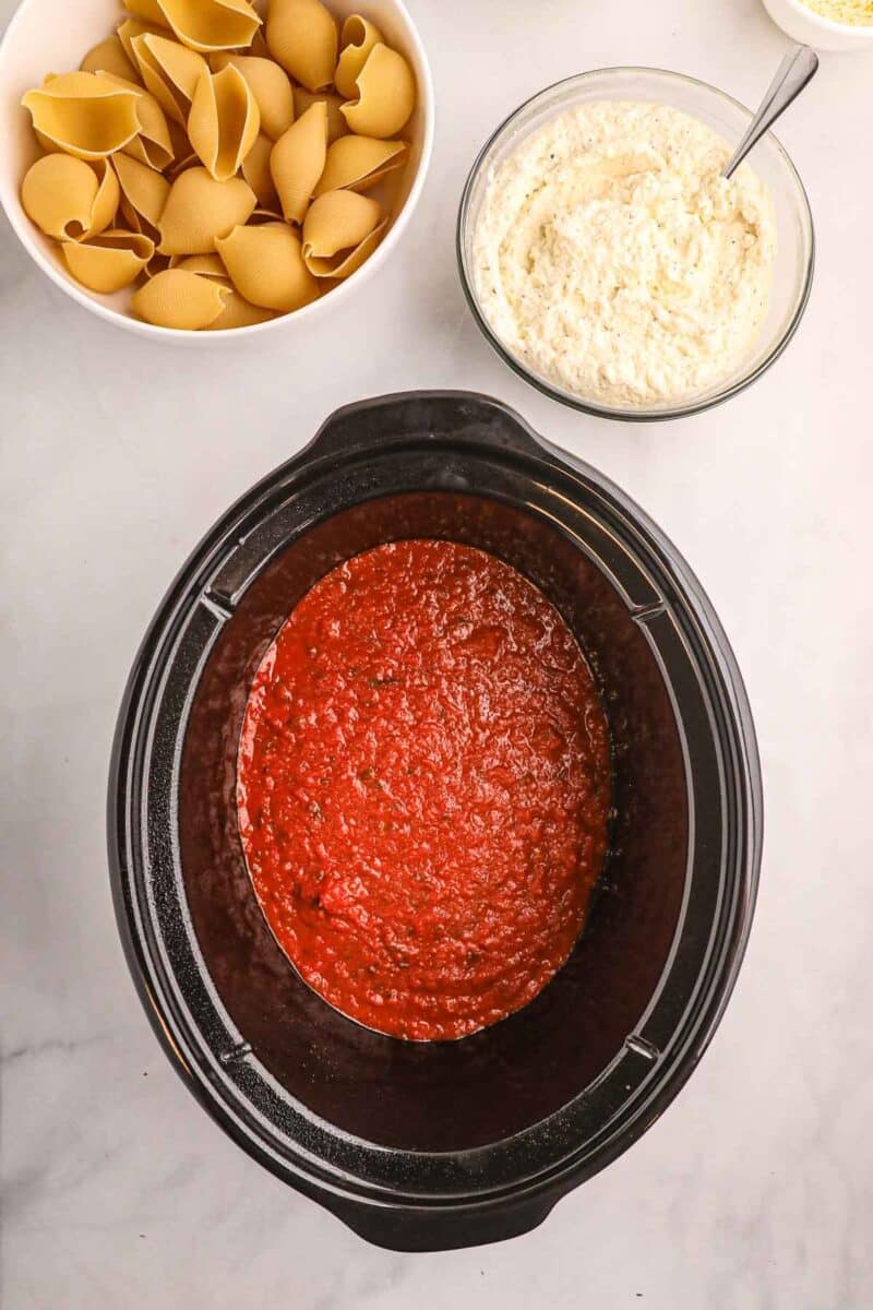 A slow cooker with tomato sauce, a bowl of uncooked pasta shells, and a bowl of ricotta cheese mixture on a white surface.