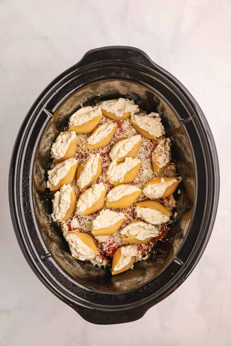 Uncooked stuffed pasta shells arranged in a slow cooker with tomato sauce and cheese visible underneath on a light countertop.