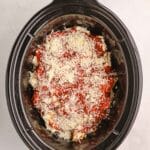 A slow cooker filled with uncooked lasagna layered with tomato sauce and shredded cheese, viewed from above on a white surface.