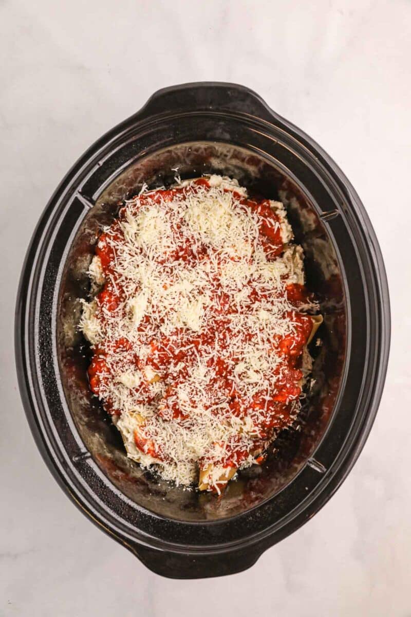 A slow cooker filled with uncooked lasagna layered with tomato sauce and shredded cheese, viewed from above on a white surface.