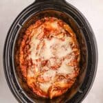 Lasagna with melted cheese and meat sauce is shown inside a black slow cooker on a light-colored surface.