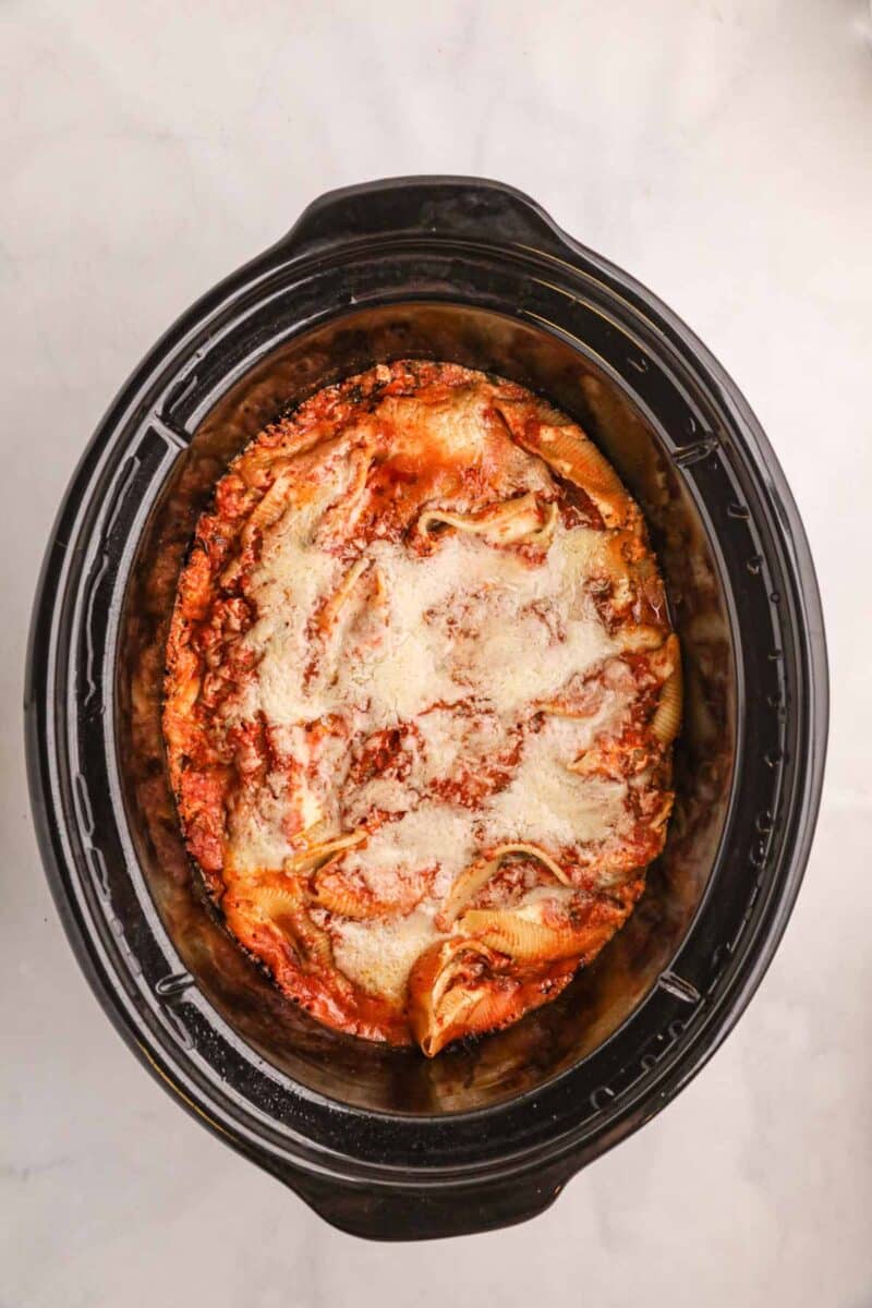Lasagna with melted cheese and meat sauce is shown inside a black slow cooker on a light-colored surface.