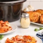 A plate of baked pasta shells with tomato sauce and cheese sits on a table beside a slow cooker, a shaker of grated cheese, and slices of garlic bread.