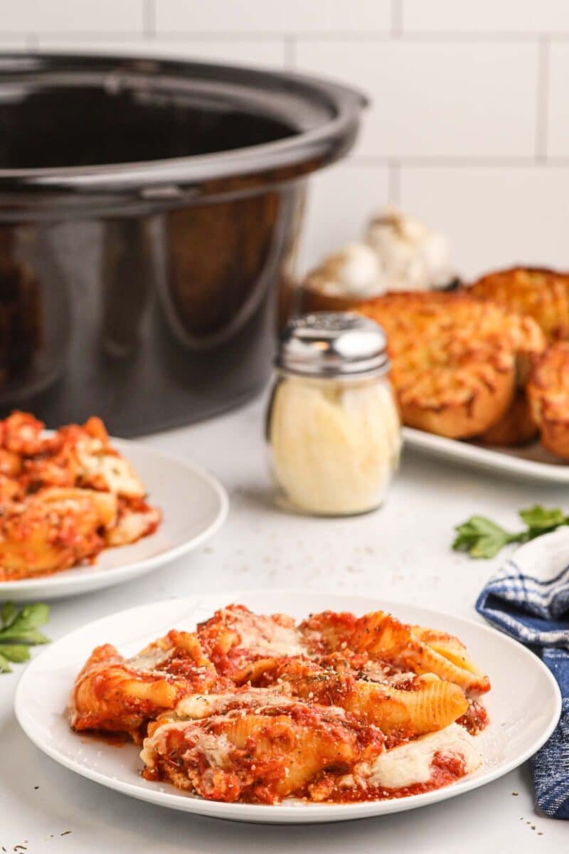 A plate of baked pasta shells with tomato sauce and cheese sits on a table beside a slow cooker, a shaker of grated cheese, and slices of garlic bread.