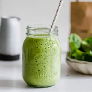 A mason jar filled with a green smoothie and a polka dot straw, set on a white surface with spinach leaves in the background.