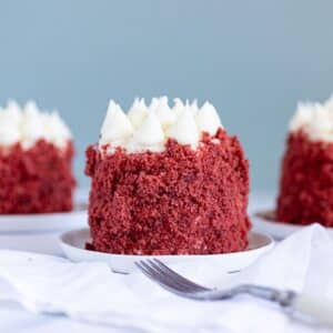 A small red velvet cake with cream cheese frosting sits on a white plate, with a fork and white napkin beside it. Two similar cakes are in the background.