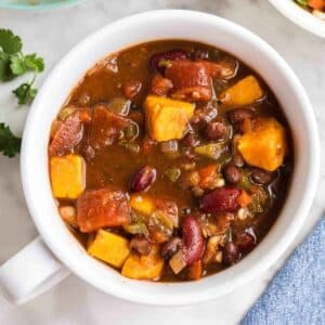 A white bowl filled with vegetable chili containing beans, tomatoes, and chunks of sweet potato on a light surface.