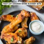 A plate of cooked chicken wings garnished with herbs, served with a side of dipping sauce, with a tray of more wings in the background. Text reads "10 Appetizers That Let Chicken Shine In Every Bite.