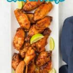 A rectangular white plate of glazed chicken wings garnished with lime wedges sits on a white surface next to a navy napkin and wooden tongs.